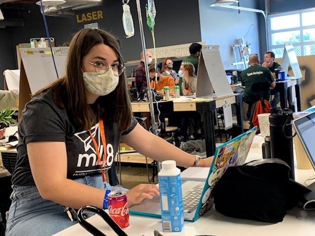 Clare pauses her CAD work to smile for the camera. Although, her smile is obscured by a thin, fabric mask.